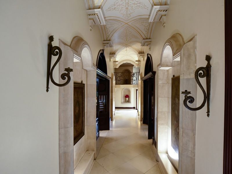 Elegant hallway featuring a vaulted ceiling with intricate decorative molding, stone flooring, and arched wall niches with integrated wrought iron sconces.