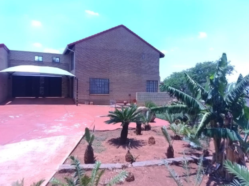 Front exterior view of a brick house featuring a paved driveway, shade sail carport, and established garden landscaping.