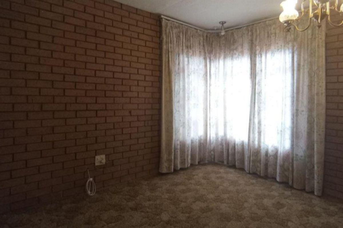 An empty living room featuring exposed brick walls, a large bay window, and carpeted flooring, offering a blank canvas for renovation.