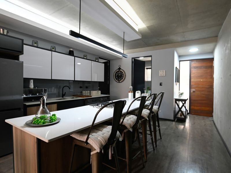Modern kitchen featuring a sleek island with quartz countertops, high-gloss cabinetry, and exposed concrete ceilings, leading to a timber entrance door.