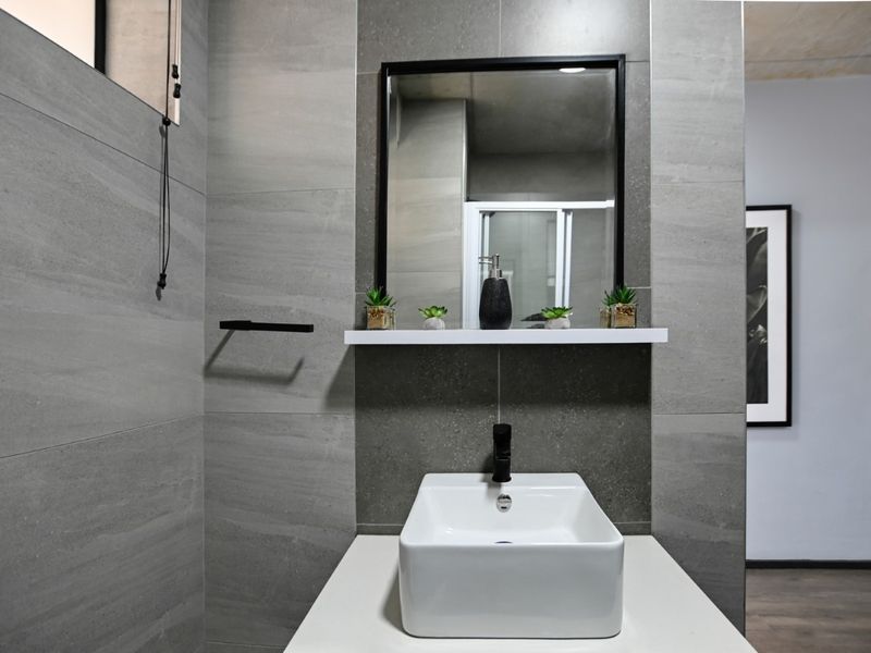 Modern bathroom featuring a sleek vessel sink, black fixtures, and contemporary grey wall tiling with a minimalist shelf and mirror.