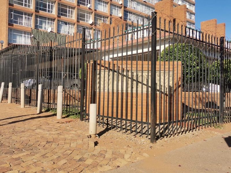 Street-level view of the apartment complex entrance featuring secure steel perimeter fencing, razor wire, and brick boundary walls with paved access.