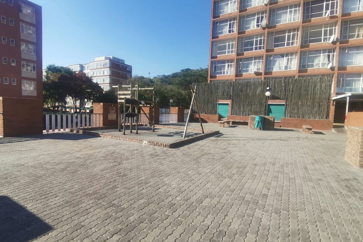 A paved communal outdoor area featuring a playground structure, brick boundary walls, and access to the apartment building complex.