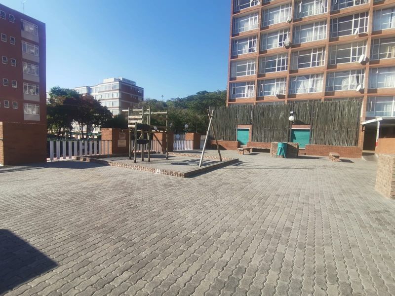 A paved communal outdoor area featuring a playground structure, brick boundary walls, and access to the apartment building complex.