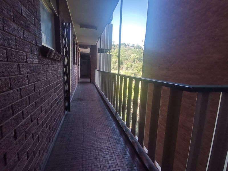 An exterior apartment walkway or balcony featuring brick walls, tiled flooring, and a metal balustrade overlooking a green landscape.