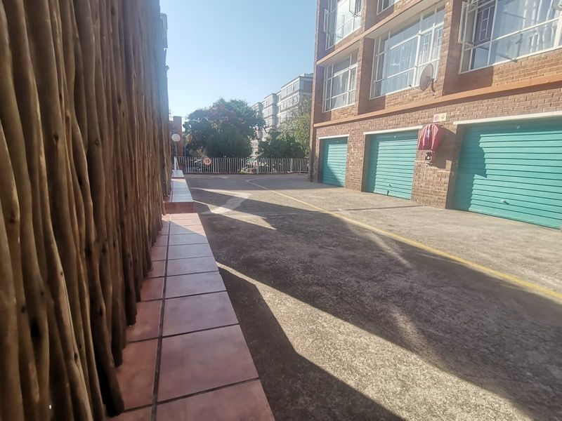 Exterior view of an apartment complex featuring a concrete driveway, multiple garage doors, and a tiled walkway with a wooden privacy screen.