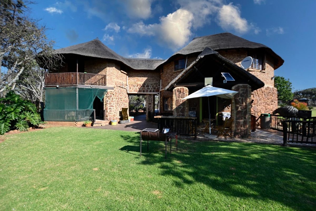 Rear view of a rustic stone-clad small holding featuring a thatched roof, covered patio area, and an upper-level balcony overlooking a well-maintained garden.
