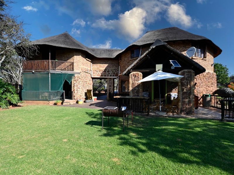 Rear view of a rustic stone-clad small holding featuring a thatched roof, covered patio area, and an upper-level balcony overlooking a well-maintained garden.