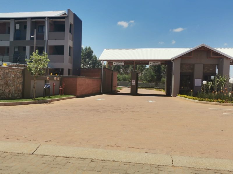 The main entrance to the apartment complex featuring a secure gatehouse, paved driveway, and modern architectural finishes.