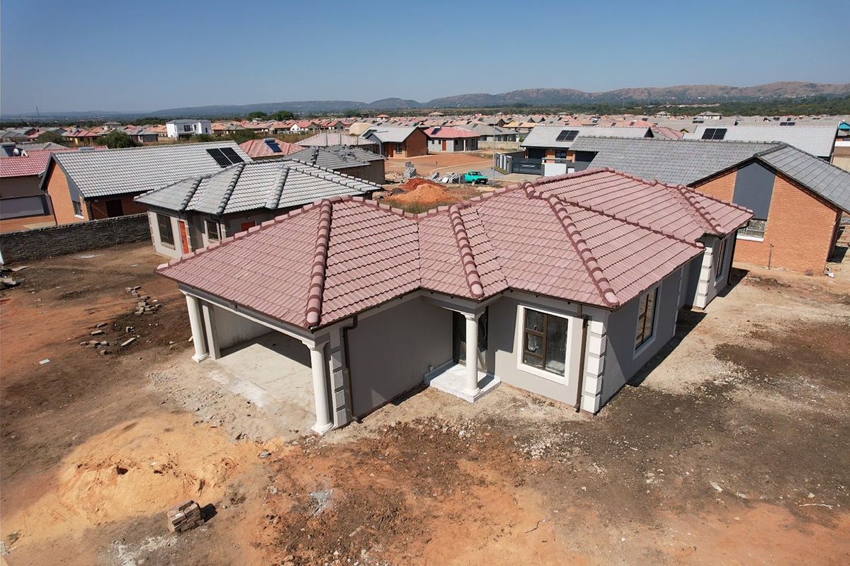 Aerial view of a newly constructed single-story house featuring a tiled roof, integrated garage, and unfinished surrounding grounds in a residential estate.