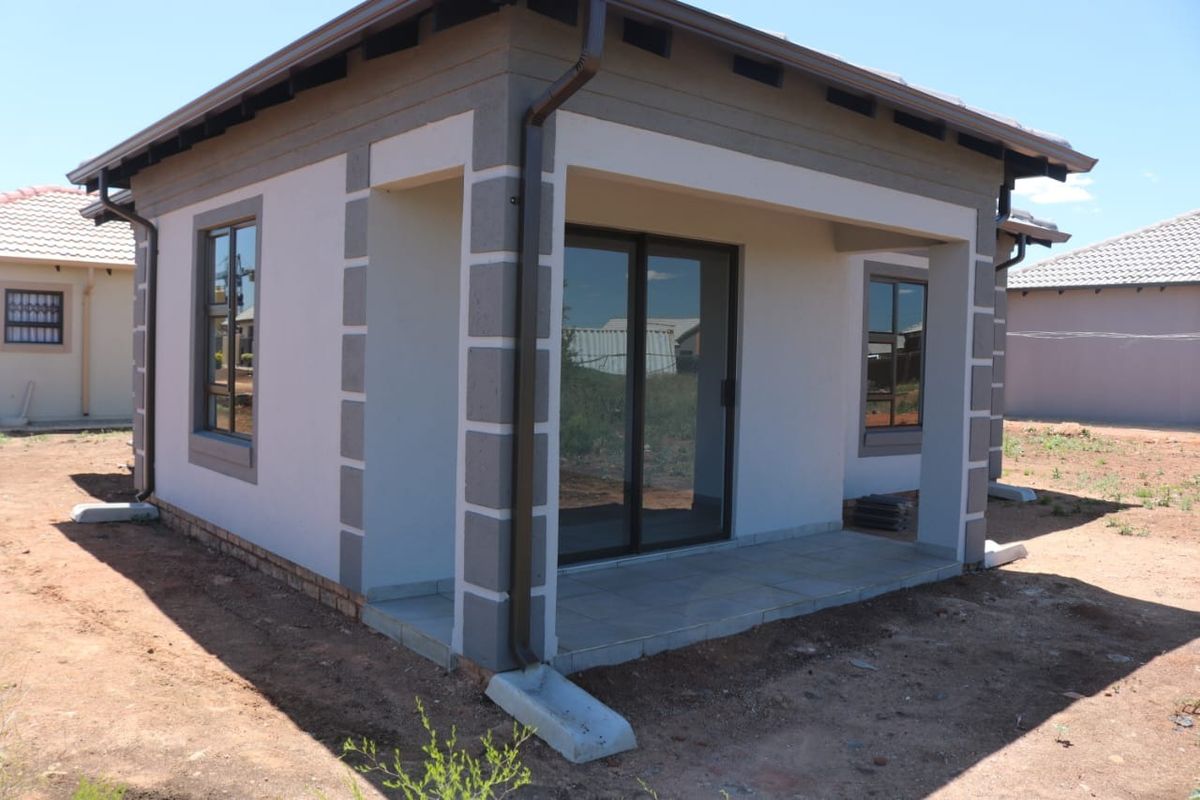 A modern residential exterior featuring a covered tiled patio, sliding glass doors, and aluminium window frames, set within an unfinished garden area.