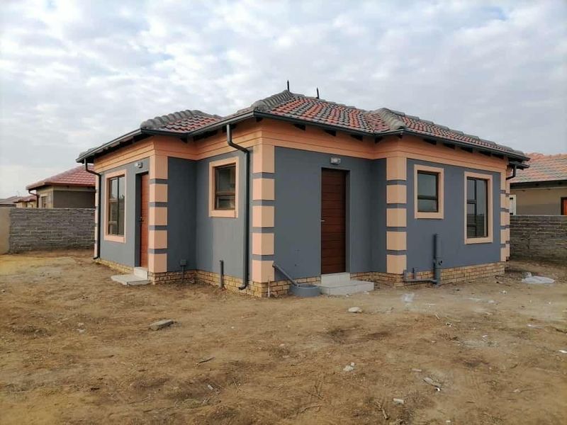A modern single-story house exterior featuring a tiled roof, grey and peach painted facade, and established window and door frames on a vacant plot.