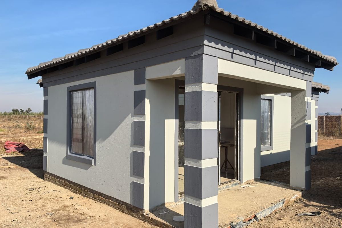 A newly constructed single-story house featuring a covered porch, tiled roof, and modern two-tone exterior paint finish, set on an undeveloped plot.