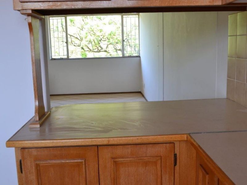 A dated kitchen area featuring wooden cabinetry and a tiled backsplash, opening into an adjacent living space with large windows.