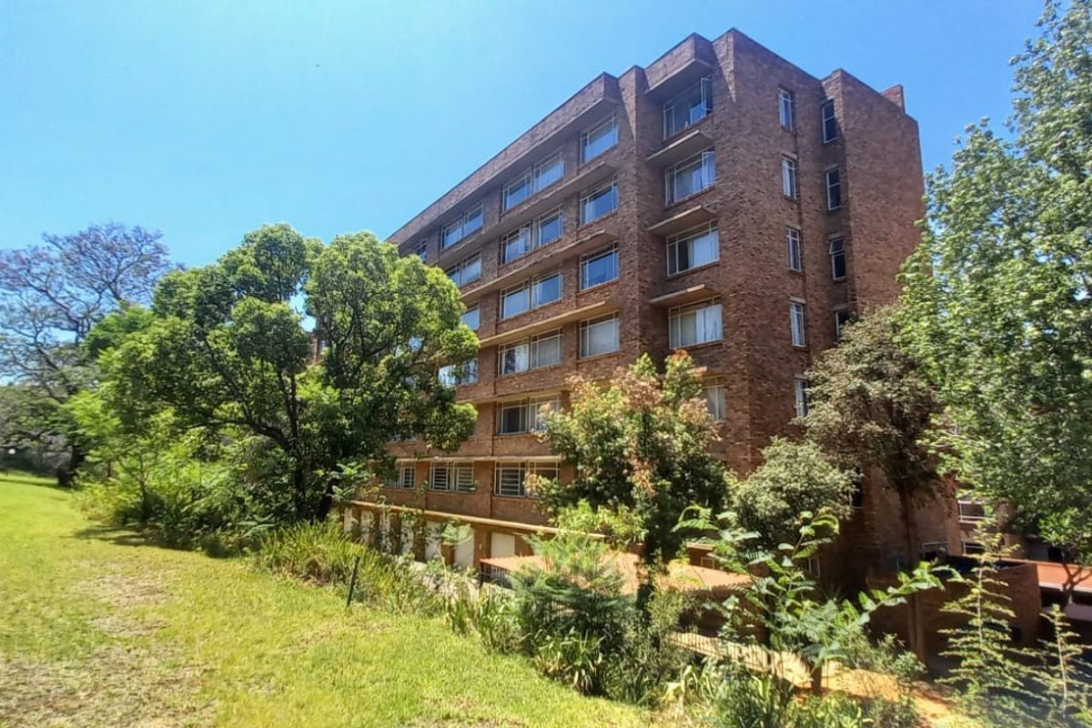A view of the rear elevation of a multi-story brick apartment building, featuring large windows and surrounding landscaped garden areas.
