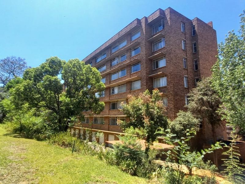 A view of the rear elevation of a multi-story brick apartment building, featuring large windows and surrounding landscaped garden areas.