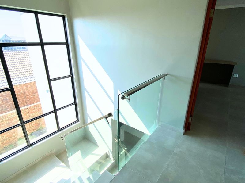 A bright, modern hallway featuring a glass balustrade with a stainless steel handrail and a large steel-framed window overlooking the exterior.