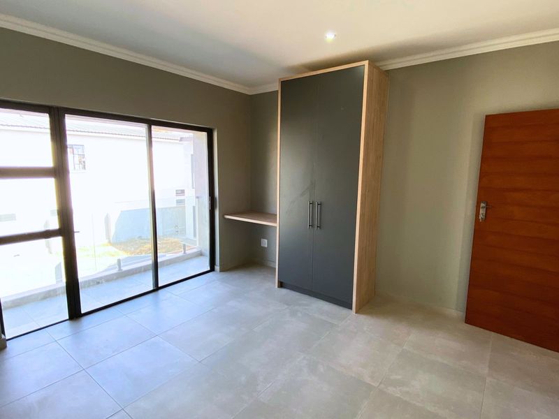 A bright, modern bedroom featuring neutral tiled flooring, a built-in wardrobe with an integrated desk, and large sliding glass doors leading to the exterior.