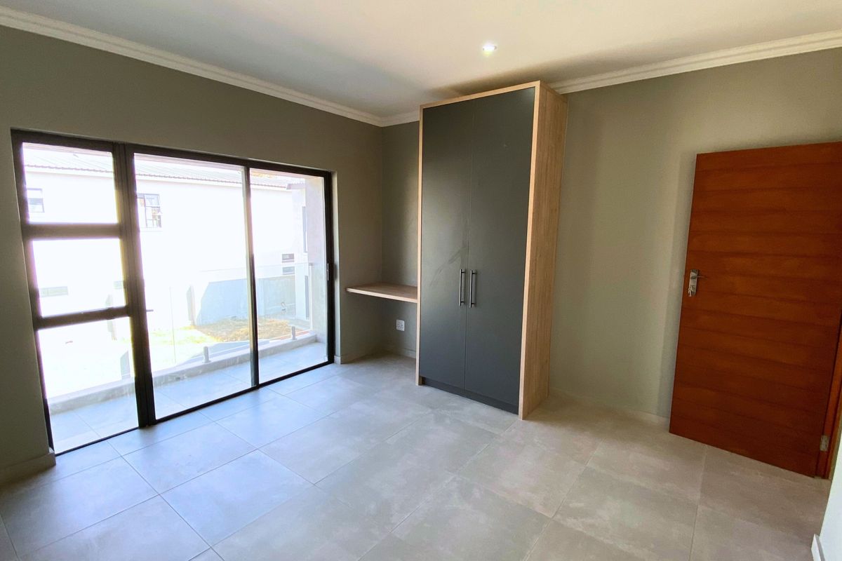 A bright, modern bedroom featuring neutral tiled flooring, a built-in wardrobe with an integrated desk, and large sliding glass doors leading to the exterior.