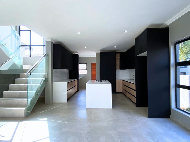 Modern open-plan kitchen featuring sleek black and wood-tone cabinetry, a central island, stone countertops, and an adjacent contemporary staircase with glass balustrades.