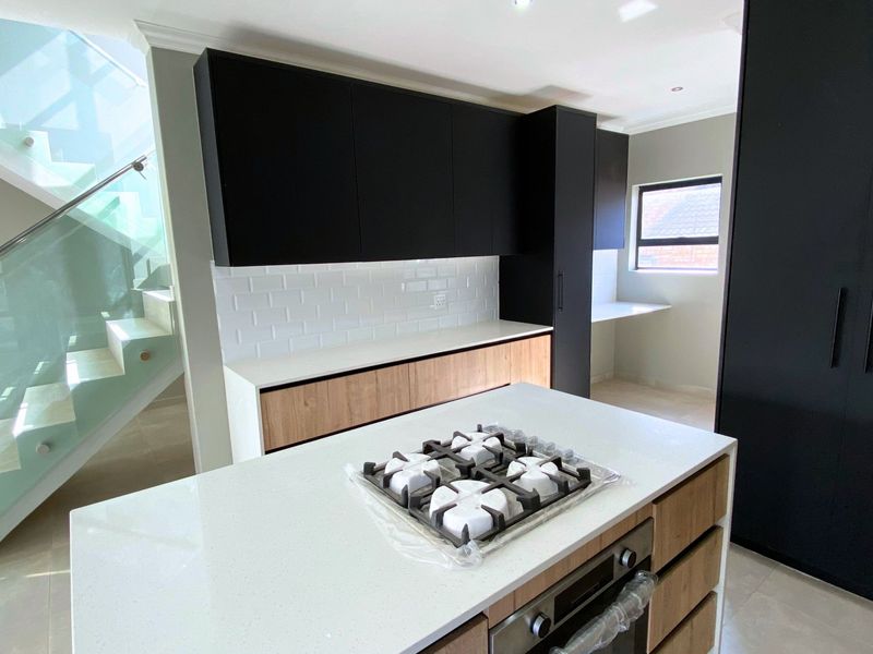 Modern kitchen featuring a central island with gas cooktop, built-in oven, sleek two-tone cabinetry, and subway tile backsplash, adjacent to a glass-railed staircase.