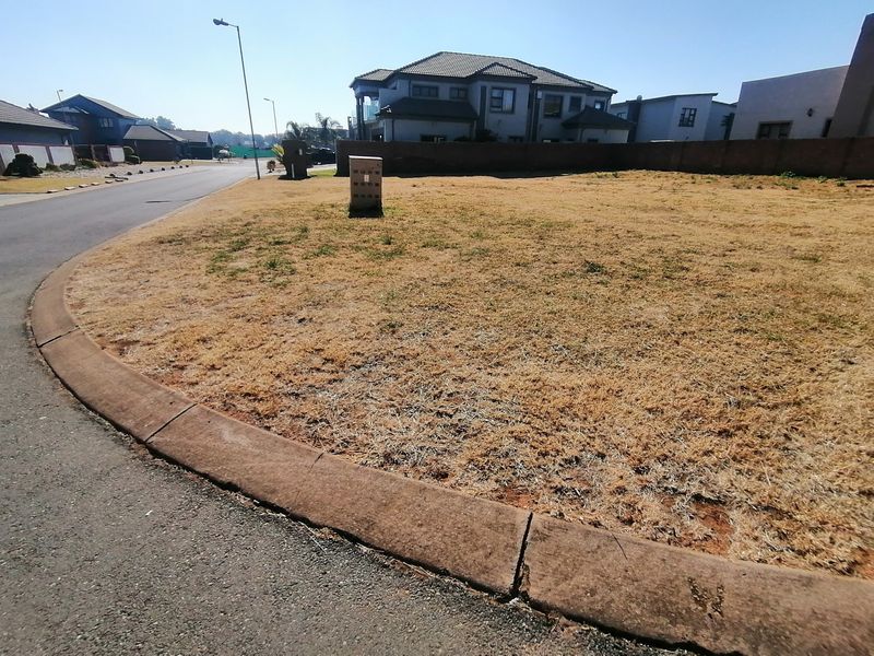 A vacant residential plot situated on a corner lot, featuring established kerbing, street lighting, and surrounding boundary walls in a developed neighborhood.