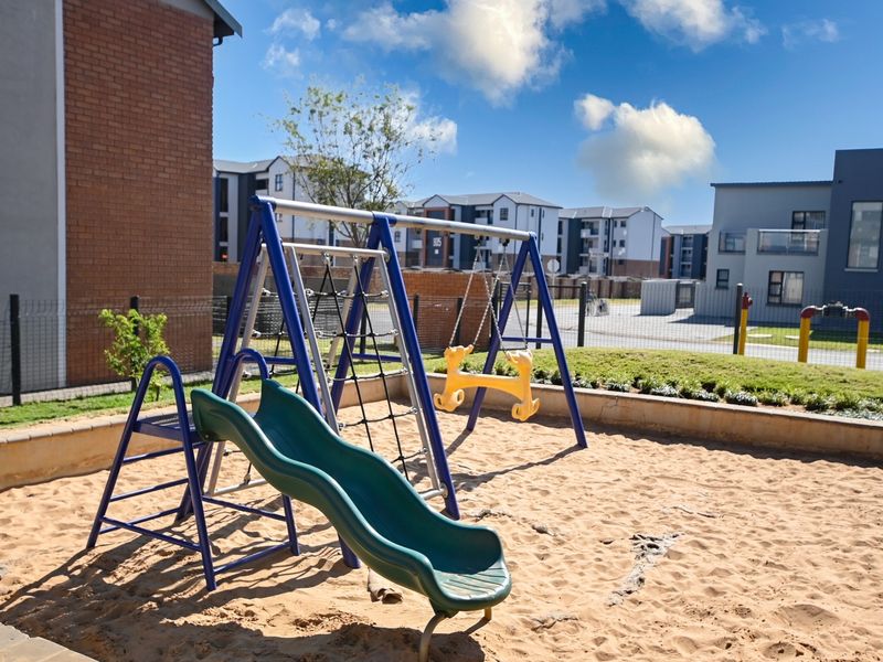 A well-maintained residential playground area featuring a sand pit, swing set, and slide, enclosed by perimeter fencing within a modern apartment complex.