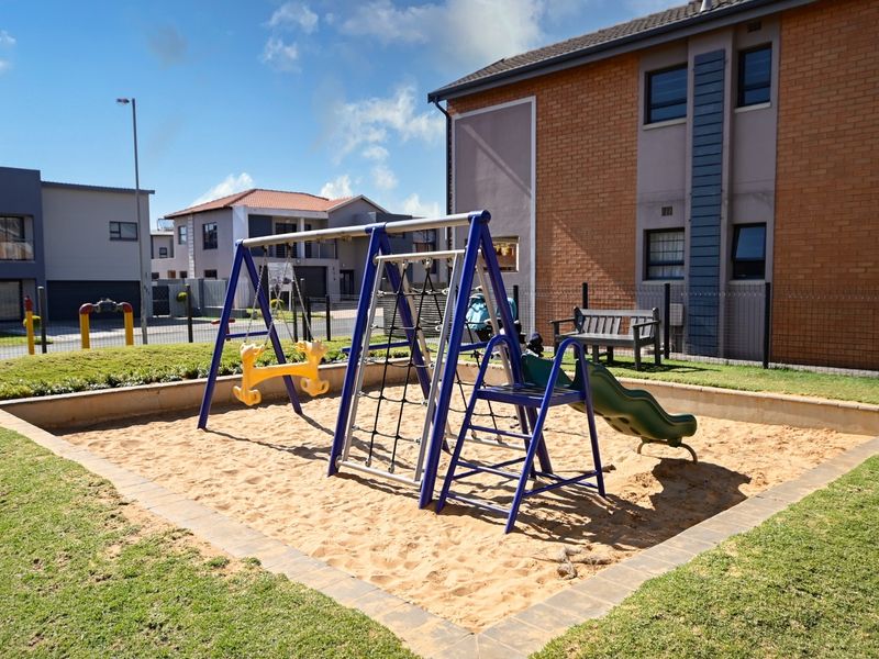 A well-maintained communal garden area featuring a sand pit with integrated playground equipment, surrounded by lawn and secure perimeter fencing.