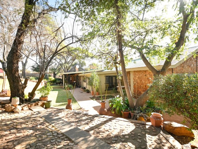 A rear view of the property featuring stone-clad exterior walls, a paved walkway, and mature trees within a landscaped garden setting.