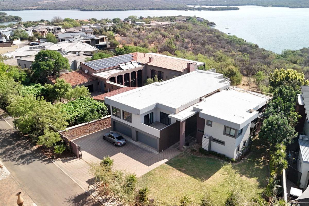 Aerial view of a modern multi-level residence featuring a flat roof, solar panels, paved driveway, and double garage, situated in a scenic lakeside estate.