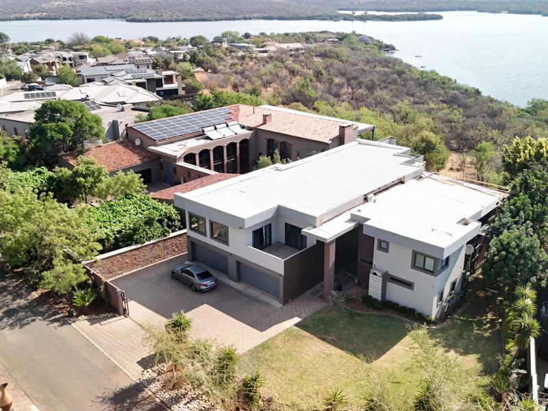 Aerial view of a modern multi-level residence featuring a flat roof, solar panels, paved driveway, and double garage, situated in a scenic lakeside estate.