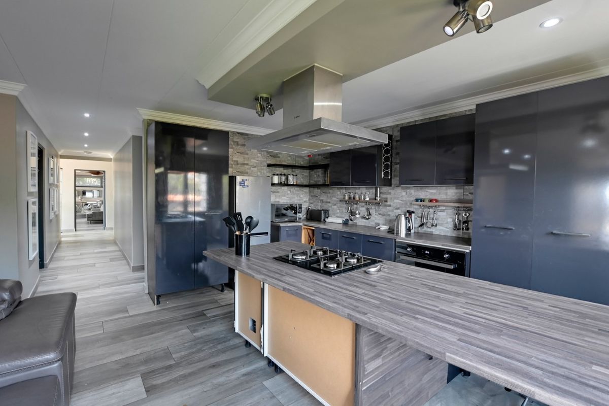 Modern kitchen featuring a central island with gas cooktop, stone veneer backsplash, dark cabinetry, and integrated appliances, leading into a hallway.