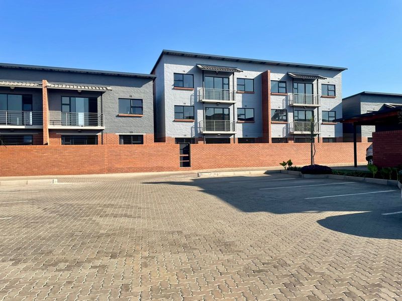 Exterior view of a modern apartment complex featuring brick boundary walls, paved parking area, and multiple balconies with metal railings.