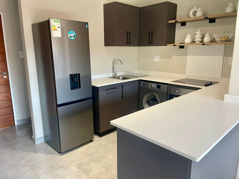 Modern kitchen featuring dark cabinetry, white quartz countertops, integrated appliances, and floating shelving, finished with neutral tiled flooring.