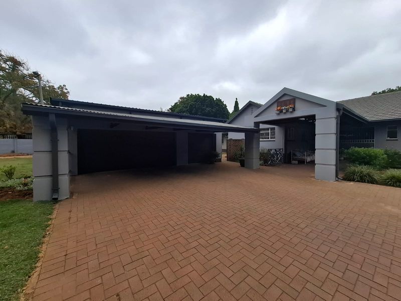 A well-maintained front exterior featuring a spacious paved driveway, a large carport, and a covered entrance area with established garden surroundings.
