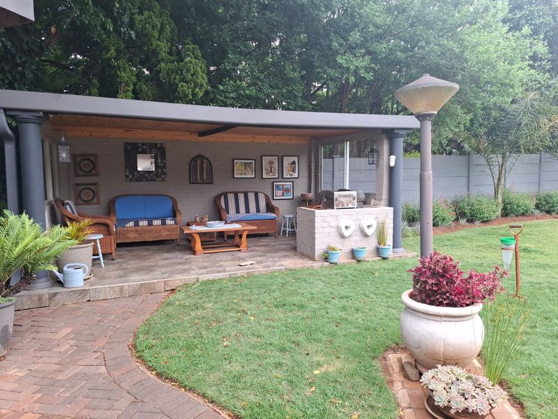 A covered outdoor patio area featuring tiled flooring, a built-in braai, and structural support columns, overlooking a landscaped garden.