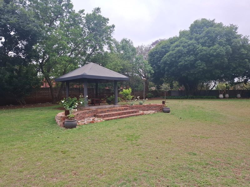 A spacious garden featuring a raised brick patio area with a covered gazebo, surrounded by mature trees and established lawn.