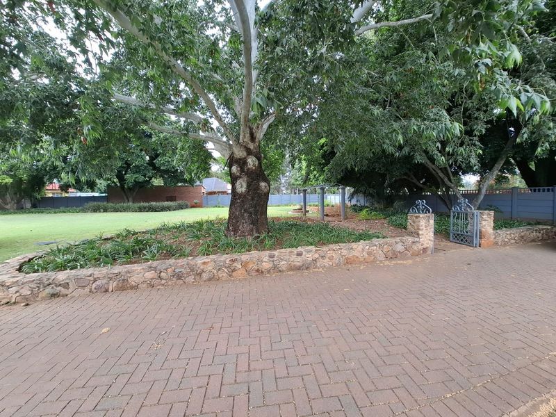 A spacious garden area featuring a paved driveway, mature trees, stone retaining walls, and a secure wrought iron gate entrance.