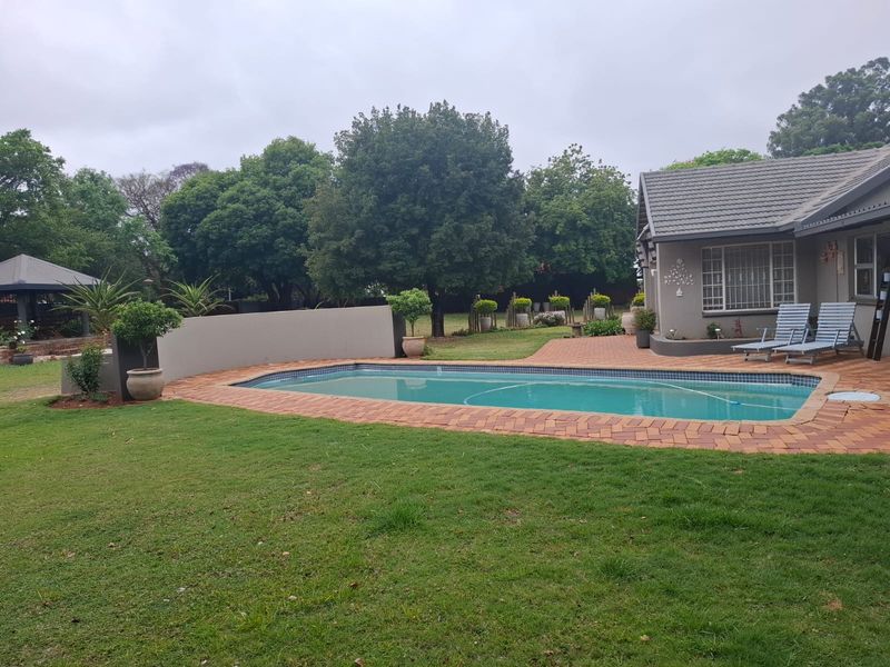 A well-maintained outdoor pool area featuring a brick-paved surround, a boundary wall, and a spacious garden setting.