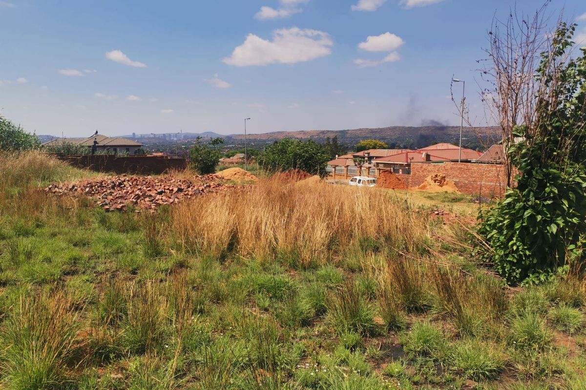 A vacant residential plot featuring natural grass, scattered construction debris, and a brick boundary wall with surrounding neighborhood views.