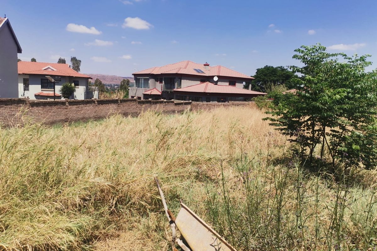 A view of a vacant residential plot featuring a brick boundary wall with electric fencing, surrounded by established residential properties.
