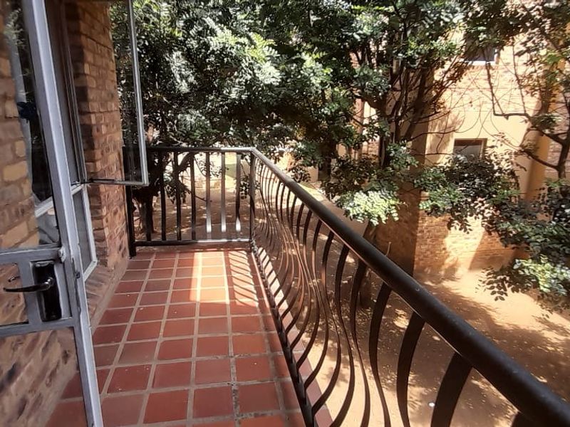 A private balcony featuring terracotta floor tiles, a metal balustrade, and an exposed brick wall, overlooking a leafy courtyard area.