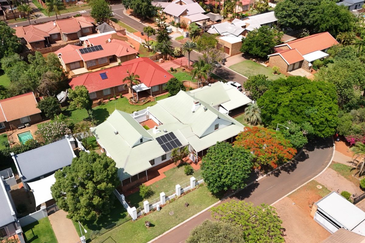 Aerial view of a residential property featuring a metal roof with solar panels, surrounded by established gardens and a paved driveway in a suburban setting.