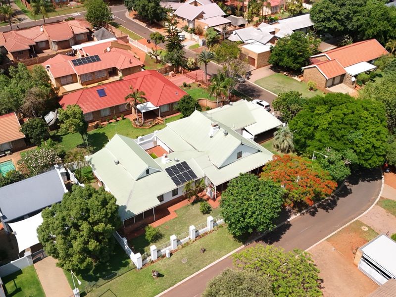 Aerial view of a residential property featuring a metal roof with solar panels, surrounded by established gardens and a paved driveway in a suburban setting.