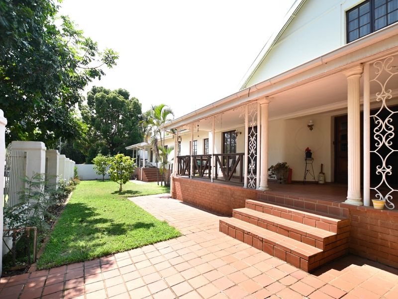 A well-maintained rear exterior featuring a covered patio with decorative columns, brick steps, and a paved walkway leading to a landscaped garden area.