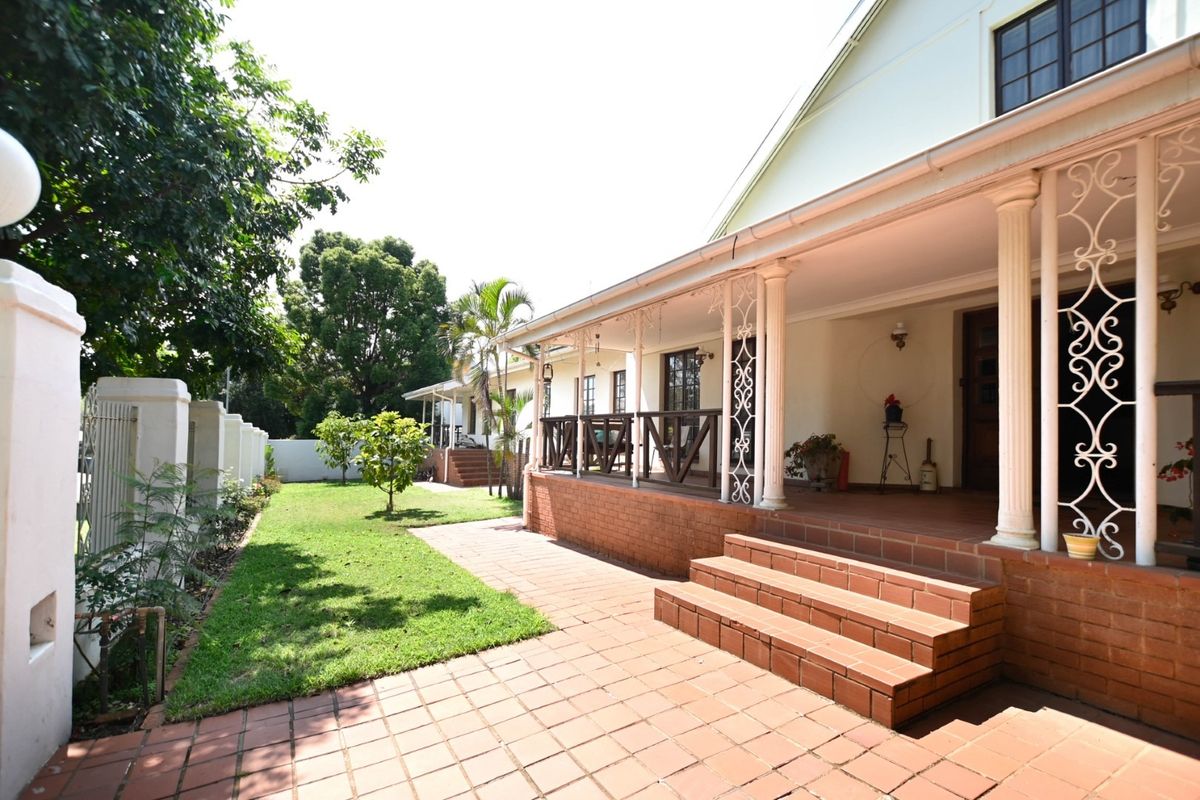 A well-maintained rear exterior featuring a covered patio with decorative columns, brick steps, and a paved walkway leading to a landscaped garden area.
