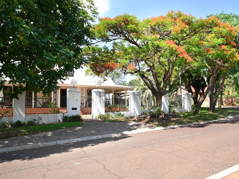 Front elevation of the property featuring a paved driveway, secure wrought iron fencing with masonry pillars, and a covered veranda surrounded by mature trees.