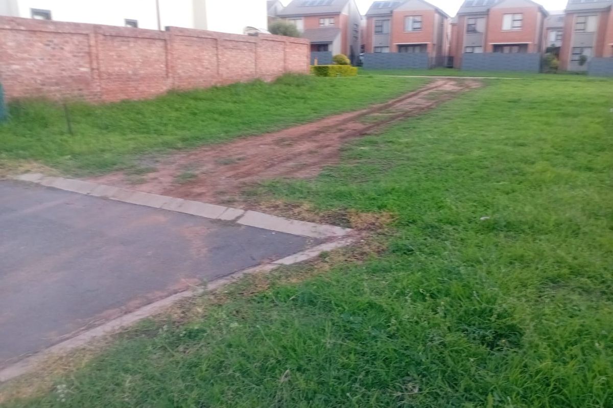 A vacant residential plot featuring a brick boundary wall, paved driveway entrance, and established grass, situated in a developed residential neighborhood.
