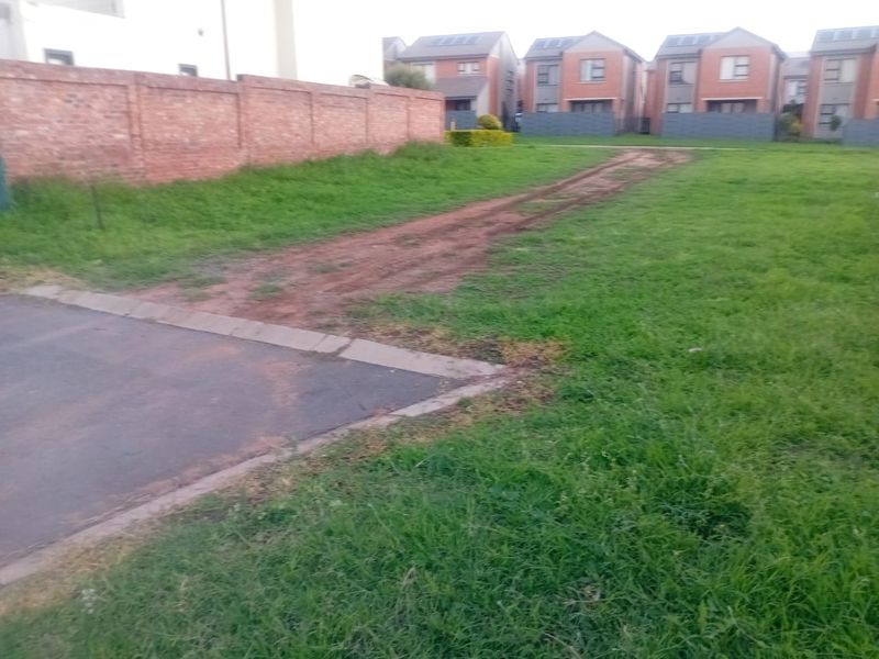 A vacant residential plot featuring a brick boundary wall, paved driveway entrance, and established grass, situated in a developed residential neighborhood.