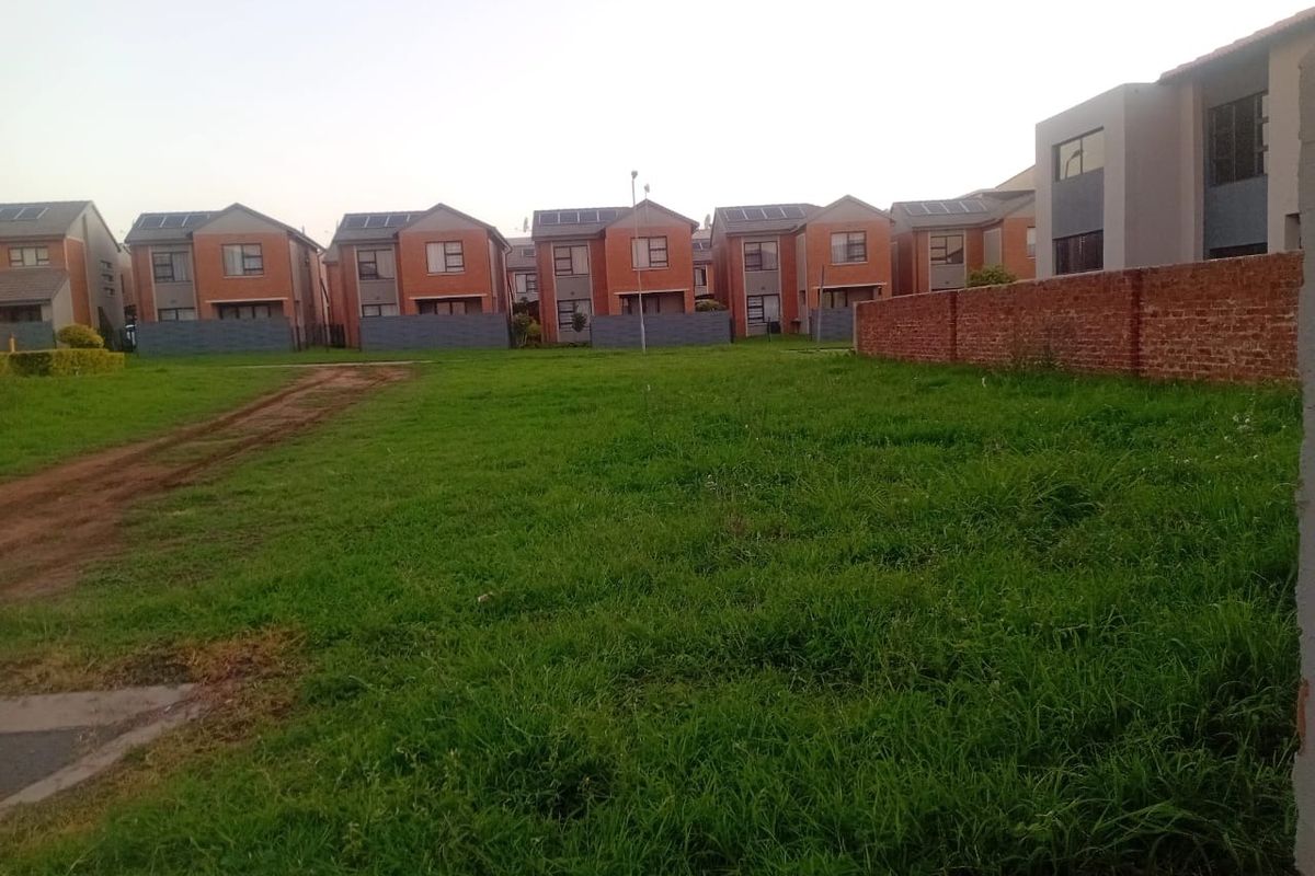 A vacant residential plot featuring a grass lawn and brick boundary wall, situated in a developed neighborhood with surrounding residential homes.